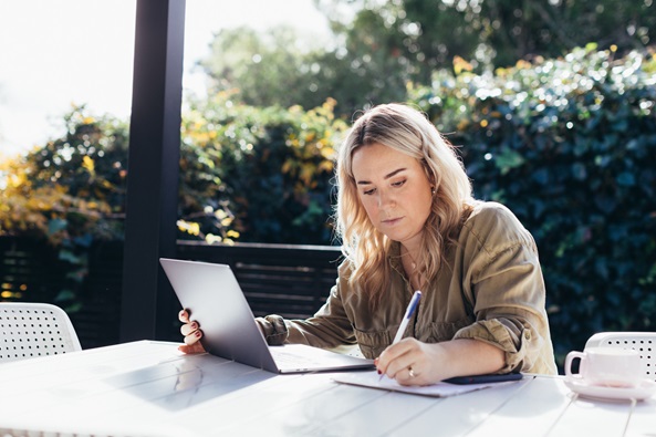 A young woman with blond here is sitting outside at a table writing notes with her laptop open.  We see trees and the sky in the background.