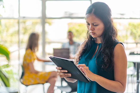 A Maori woman with long dark hair is standing and viewing her iPad. A woman and man are seated at a table behind her