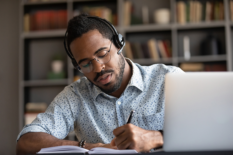 Man with dark hair and skin, wearing headphones is studying in a library